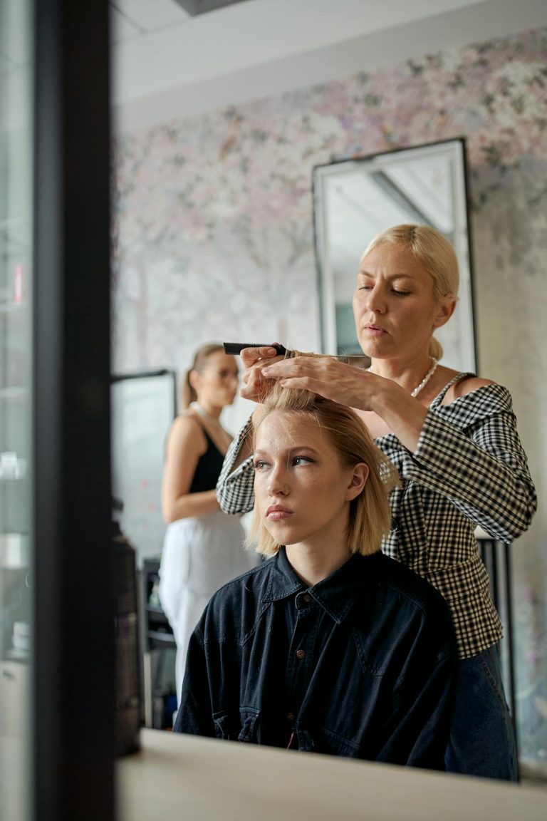 Adult female hairstylist straightening hair of dreamy client with iron reflecting in mirror in beauty salon