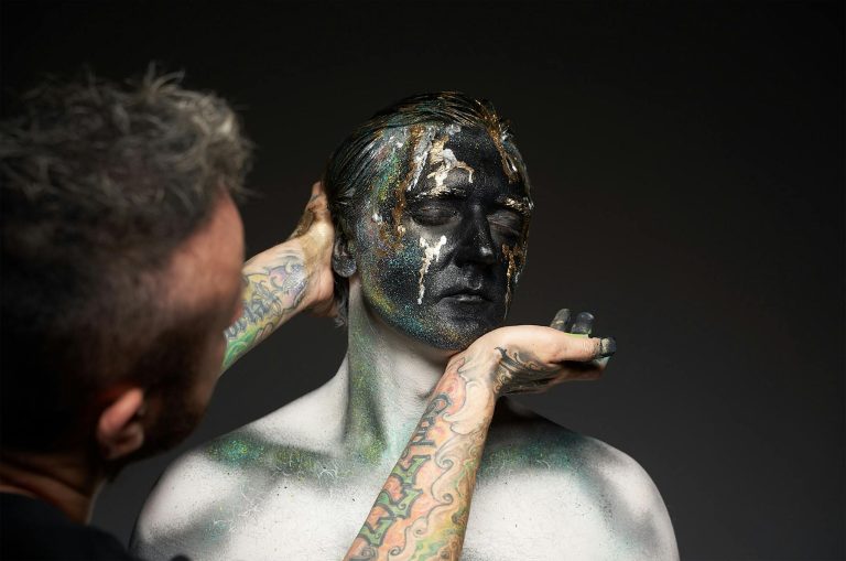 Side view of crop anonymous man applying artistic makeup on face of male model during creative photo session against black background